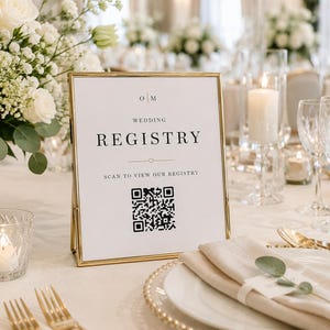 May include: A rectangular wedding registry sign with a gold metal frame, featuring black text that reads "WEDDING REGISTRY" and "SCAN TO VIEW OUR REGISTRY" above a black and white QR code, with the initials "O M" printed at the top center.
