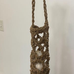 May include: A brown, macrame-style garlic holder with a woven design. The holder is suspended by a rope and contains several white garlic bulbs. The natural fiber construction gives it a rustic, handmade appearance.