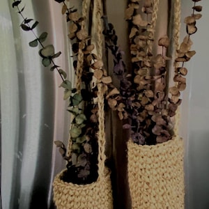 May include: Two cylindrical, hand-crocheted beige jute holders suspended by matching loops. Each holder contains dried eucalyptus branches with brown and muted green leaves. The items are hanging from two circular silver-toned magnetic hooks.