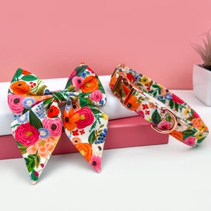 May include: A floral patterned dog collar and matching bow tie. The collar is rose gold with a white background and the bow tie is a white background with a floral pattern in shades of pink, red, orange, and green.