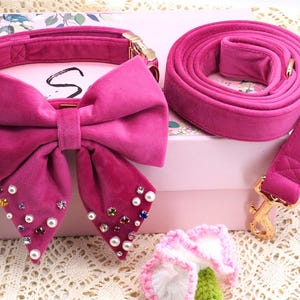 May include: A fuchsia velvet dog collar with a large bow adorned with pearls and gemstones, paired with a matching leash. The set includes a gold-tone clasp and is presented on a pink box.