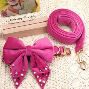 May include: A hot pink velvet dog collar with a matching leash. The collar features a large bow adorned with faux pearls and colorful rhinestones. The leash has a gold-colored clasp and hardware.