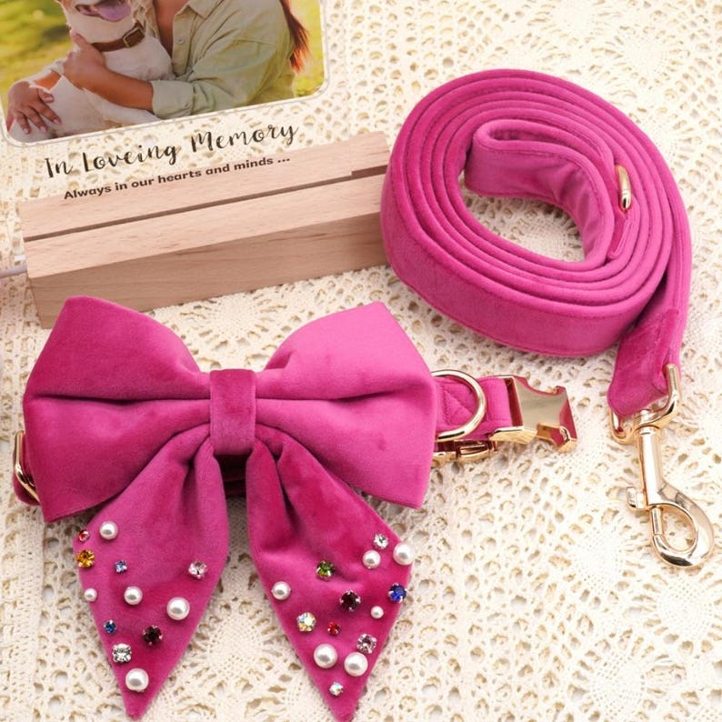 May include: A hot pink velvet dog collar with a matching leash. The collar features a large bow adorned with faux pearls and colorful rhinestones. The leash has a gold-colored clasp and hardware.