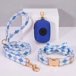 May include: A dog accessory set featuring a blue and white argyle patterned leash, collar with bow tie, and a matching waste bag dispenser. The set includes gold-colored metal hardware and a blue waste bag dispenser with a black flower design.