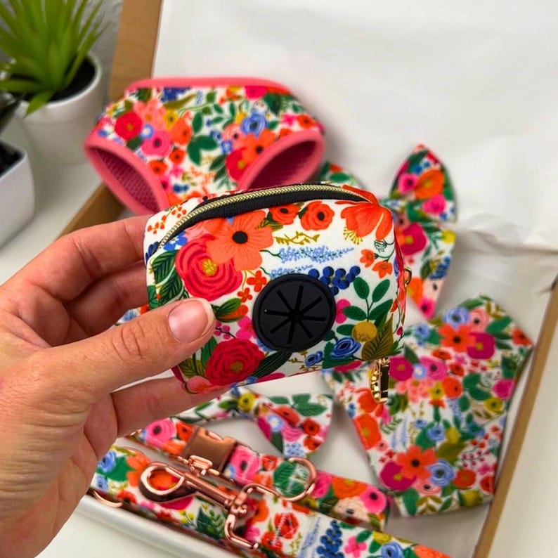 Puede incluir: Un dispensador de bolsas para excrementos de perro con estampado floral y abertura de goma negra, sostenido en una mano. El dispensador forma parte de un conjunto que incluye un arn&eacute;s, una pajarita, una correa y guantes a juego, todos con un estampado floral vibrante.