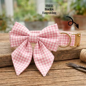 May include: A pink and white gingham patterned pet collar featuring a large, matching fabric bow attached to the front. The collar includes a gold-toned metal buckle and a D-ring for leash attachment. Text on the image reads FREE Buckle Engraving.