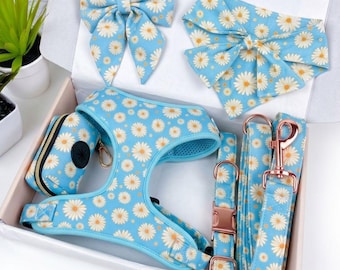 May include: A dog accessory set featuring a light blue daisy print. The set includes a harness, leash, collar, bow tie, bandana, and waste bag dispenser. The metal hardware is rose gold. The items are arranged in a white box.