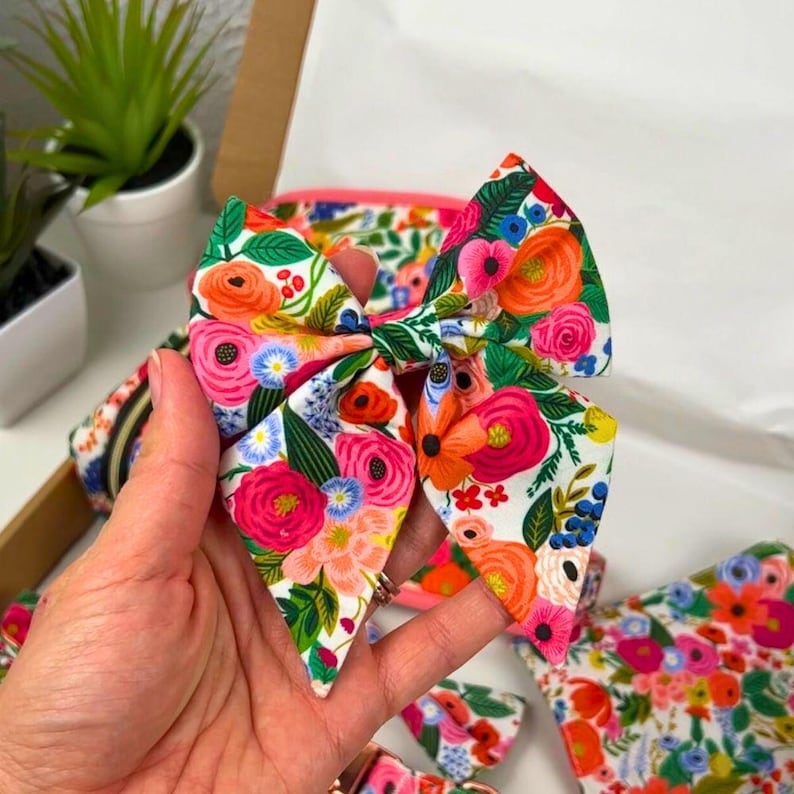 Puede incluir: Un lazo grande para el cabello de tela con un vibrante estampado floral que presenta flores rosas, naranjas y azules sobre un fondo blanco. El lazo se sostiene en una mano, con otros accesorios a juego visibles en el fondo.