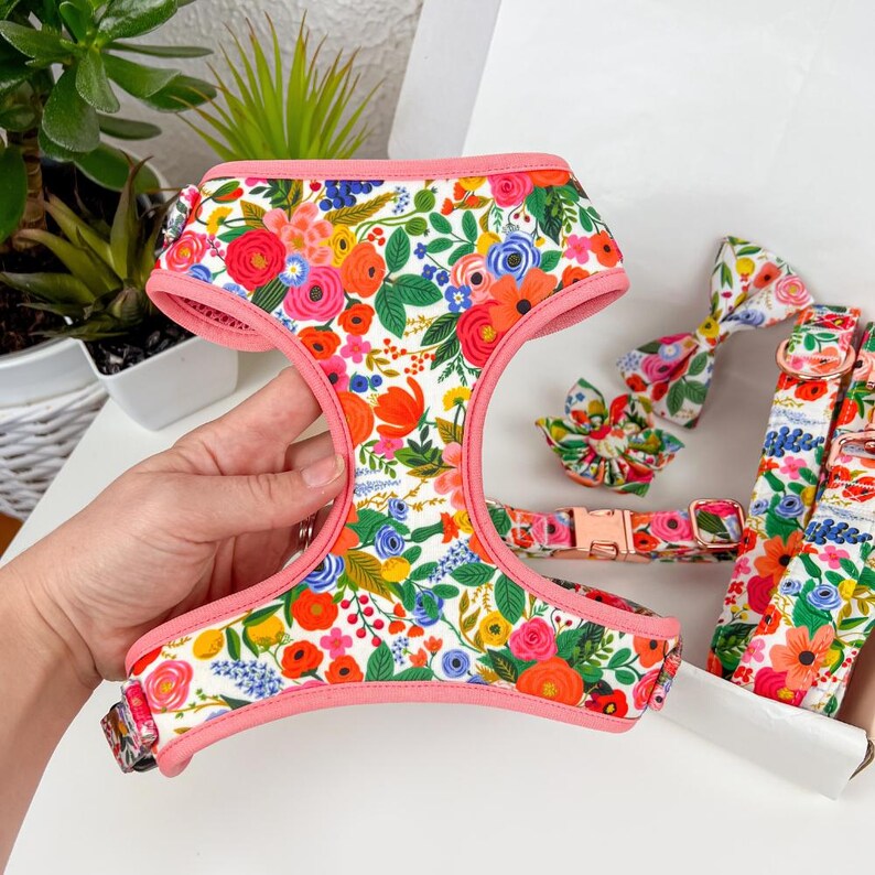 Puede incluir: Un arn&eacute;s floral para perros con pajarita y collar a juego. El arn&eacute;s, la pajarita y el collar presentan un fondo blanco con un colorido estampado floral de flores rojas, naranjas, azules y amarillas. El arn&eacute;s tiene un ribete rosa.