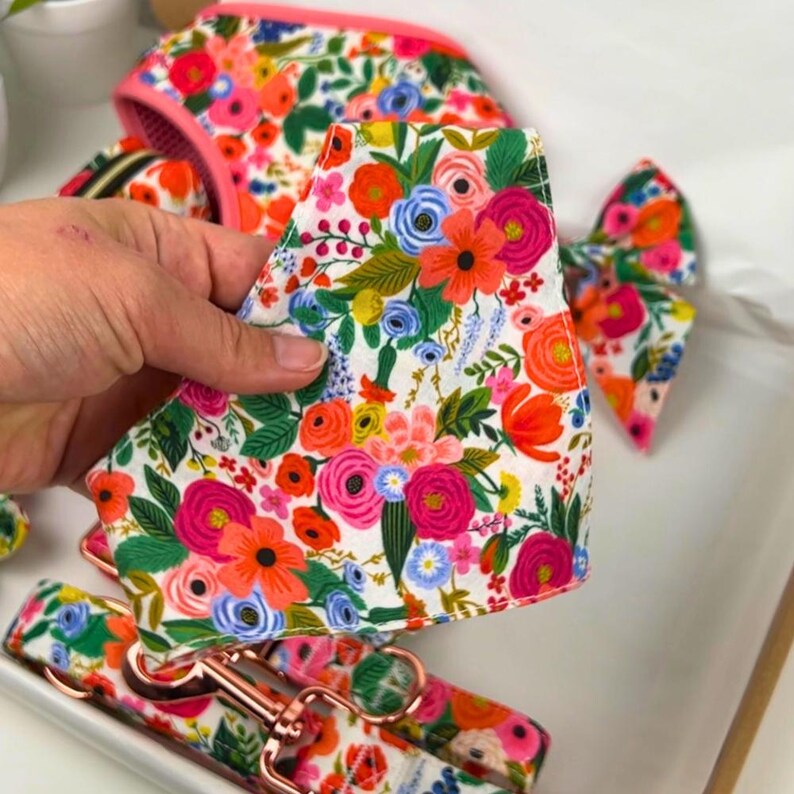Puede incluir: Un conjunto de accesorios para perros con estampado floral, que incluye un arn&eacute;s, una correa, una pajarita y una bandana. Los art&iacute;culos presentan un fondo blanco con flores vibrantes en tonos rosa, naranja, azul y verde, acentuados con herrajes de oro rosa.