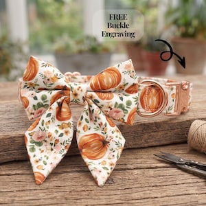 May include: A pet collar featuring a white fabric base with a repeating pattern of orange pumpkins, pink flowers, and green leaves. The collar includes a large matching bow, a rose gold metal buckle, and a D-ring. Text reads FREE Buckle Engraving.