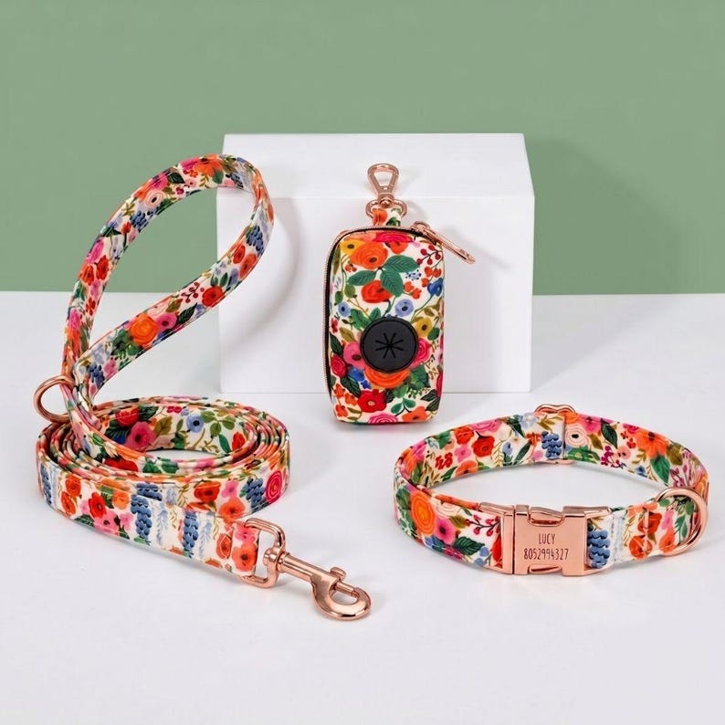 Puede incluir: Un conjunto de collar, correa y dispensador de bolsas para excrementos de perro con estampado floral. El conjunto presenta un colorido patr&oacute;n de flores en tonos naranja, rosa y azul sobre un fondo blanco. El collar tiene una hebilla de oro rosa con el nombre "LUCY" y un n&uacute;mero de tel&eacute;fono.