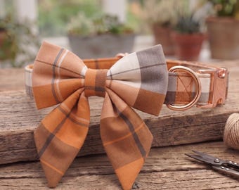 May include: A pet collar featuring a large, detachable bow tie in a burnt orange, cream, and grey herringbone plaid pattern. The collar includes rose gold metal hardware, a D-ring for leash attachment, and a matching plaid fabric strap design.