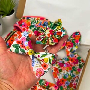Puede incluir: Una colecci&oacute;n de accesorios para mascotas con estampado floral, que incluye una pajarita, una flor y un arn&eacute;s. Los art&iacute;culos presentan una vibrante mezcla de flores rosas, naranjas, amarillas y verdes sobre un fondo blanco. Los accesorios se muestran en una caja de cart&oacute;n.