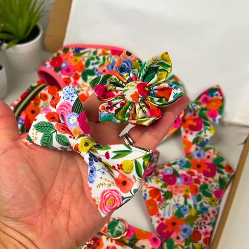 Puede incluir: Una colecci&oacute;n de accesorios para mascotas con estampado floral, que incluye una pajarita, una flor y un arn&eacute;s. Los art&iacute;culos presentan una vibrante mezcla de flores rosas, naranjas, amarillas y verdes sobre un fondo blanco. Los accesorios se muestran en una caja de cart&oacute;n.