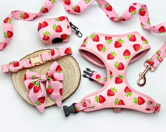 May include: Pink dog harness, collar, and leash set with a strawberry print. The set includes a bow tie, poop bag holder, and a leash with a gold metal clip.