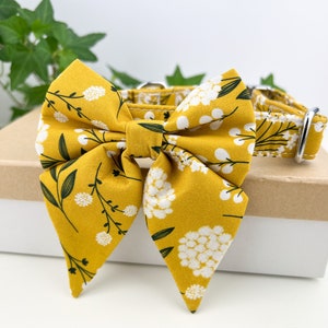 May include: A yellow dog collar with a white and green floral print bow tie. The bow tie is tied in a large, floppy bow.