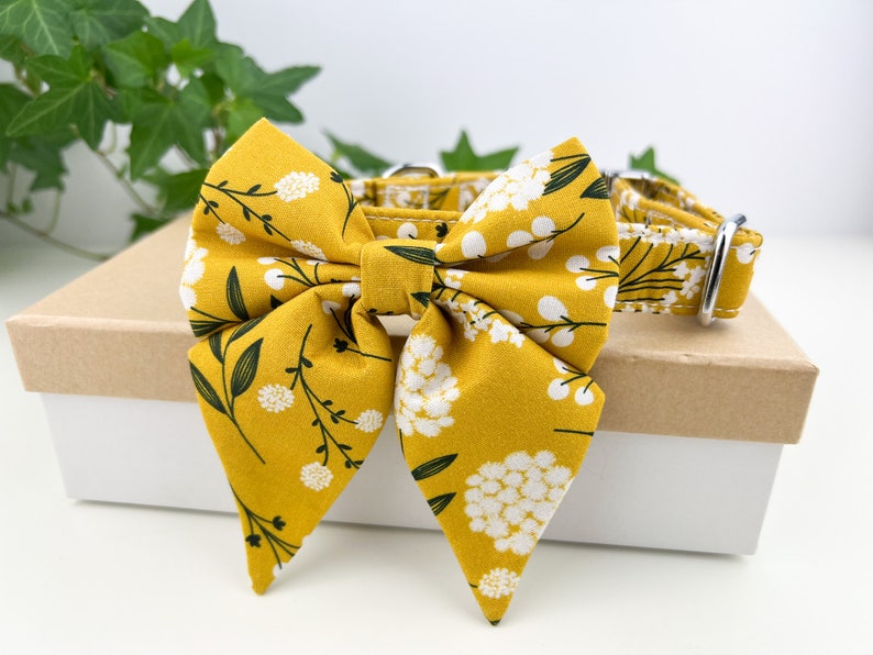 May include: A yellow dog collar with a white and green floral print bow tie. The bow tie is tied in a large, floppy bow.