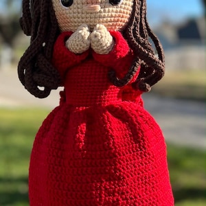 May include: A handmade crochet doll with a red dress, brown hair, and a white and red headpiece. The doll has a tan face with black eyes and is holding its hands together. The doll is a unique, handcrafted item.