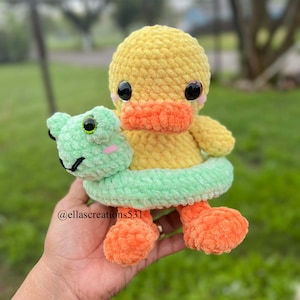 May include: A crocheted yellow duck wearing a green and yellow pool float. The duck has orange feet and black eyes. The duck is holding a green crocheted frog in its beak. The duck is sitting on a hand.