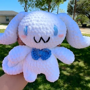 May include: A white plush toy made of soft yarn with long floppy ears, blue embroidered eyes, pink cheeks, and a black stitched mouth. The character features a small blue crocheted bow tie at the neck and has a rounded, stuffed body shape.