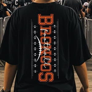 Puede incluir: Camiseta negra con la palabra "BRONCOS" en naranja y blanco, un gráfico de fútbol americano y marcadores de yardas. La camiseta la lleva una persona en un partido de fútbol americano, con un estadio lleno de espectadores al fondo.