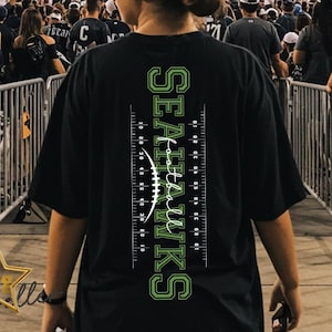 May include: Black t-shirt with the text "Seahawks" in green, along with a football field design. The shirt is worn by a person in a stadium setting, surrounded by other fans. The image captures a game day atmosphere.