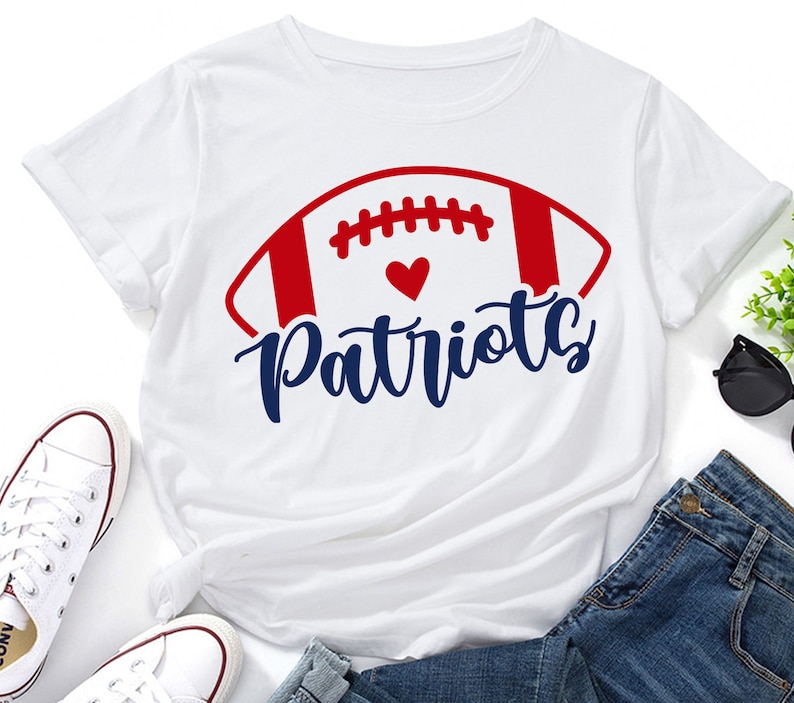 May include: White t-shirt with a red, white, and blue football graphic and the text "Patriots" in blue. A small red heart is inside the football graphic.