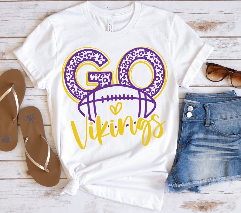 May include: White t-shirt with a graphic design that says "GO Vikings". The word "GO" is in purple leopard print, and the word "Vikings" is in yellow script. A purple football with a heart is in the center.