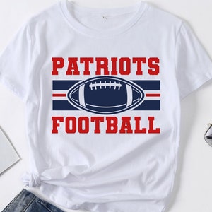 May include: White t-shirt with the words "PATRIOTS FOOTBALL" in red block letters. A navy blue football graphic is centered between the words, with a red stripe above and below the football. The shirt is made of a soft material.
