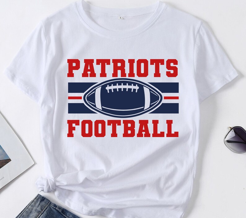 May include: White t-shirt with the words "PATRIOTS FOOTBALL" in red block letters. A navy blue football graphic is centered between the words, with a red stripe above and below the football. The shirt is made of a soft material.