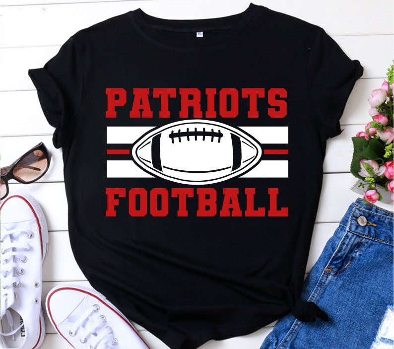 May include: Black t-shirt with the words "PATRIOTS FOOTBALL" in red block letters. A white football graphic is centered between the words, with a red stripe above and below it. The shirt is a casual style.