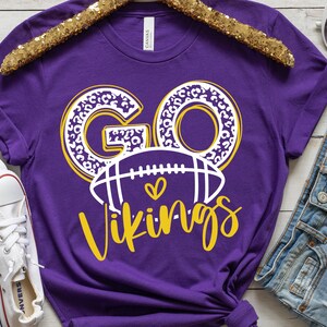 May include: Purple t-shirt with the words "GO Vikings" in a white and yellow design. The word "GO" has a leopard print pattern. A football graphic with a heart is below the word "GO".