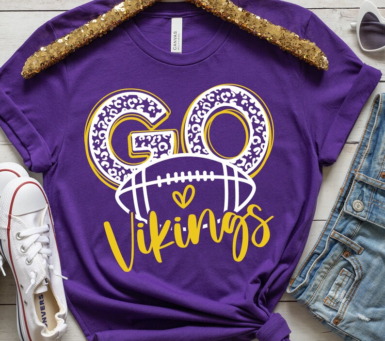 May include: Purple t-shirt with the words "GO Vikings" in a white and yellow design. The word "GO" has a leopard print pattern. A football graphic with a heart is below the word "GO".