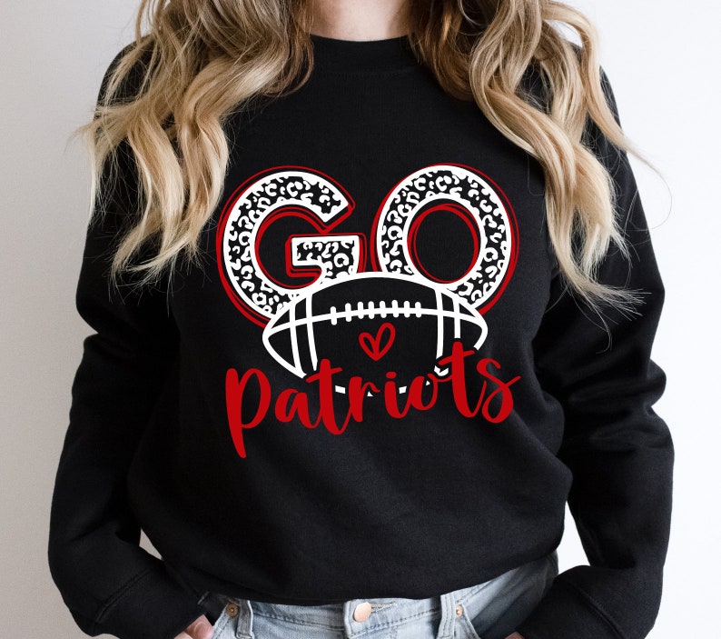 Op de afbeelding: Zwart sweatshirt met de woorden "GO Patriots" in rood en wit. Het woord "GO" heeft een luipaardprint ontwerp. Een witte voetbal met een rood hart staat tussen de woorden "GO" en "Patriots".