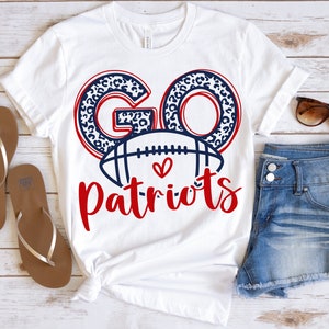 Op de afbeelding: Wit T-shirt met de woorden "GO Patriots" in rood en marineblauw. De letters "GO" hebben een luipaardprint. Een voetbalafbeelding is gecentreerd onder de woorden, met een hartsymbool. Het shirt wordt gecombineerd met een spijkerbroek en een zonnebril.