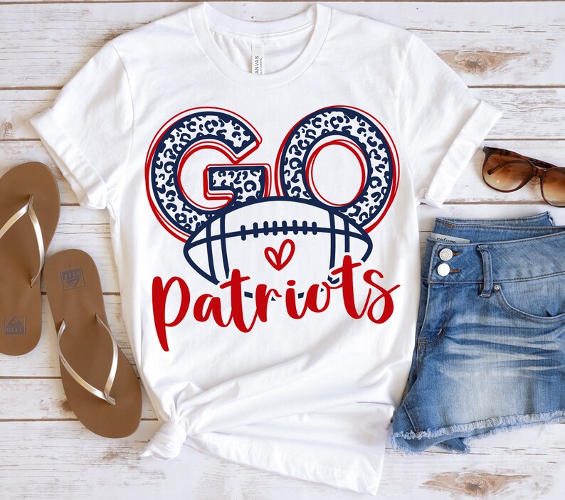 Op de afbeelding: Wit T-shirt met de woorden "GO Patriots" in rood en marineblauw. De letters "GO" hebben een luipaardprint. Een voetbalafbeelding is gecentreerd onder de woorden, met een hartsymbool. Het shirt wordt gecombineerd met een spijkerbroek en een zonnebril.