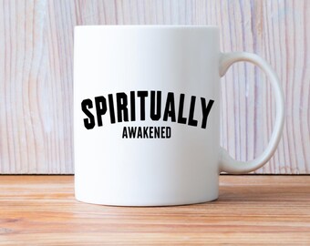 May include: A white ceramic mug with the text "SPIRITUALLY AWAKENED" in black. The mug has a curved handle and is on a wooden surface. The mug is a standard size for tea or coffee.