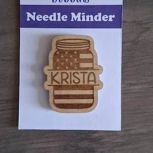 May include: A wooden needle minder shaped like a mason jar with an engraved American flag pattern and the name KRISTA centred on a horizontal bar. The item is attached to a white display card labelled Barnwood & Bobbins Needle Minder.