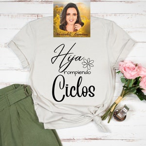 May include: White t-shirt with black text that reads "Hija rompiendo Ciclos" with a small flower graphic. The shirt is laid flat on a white wooden surface with a bouquet of pink roses and a green skirt.