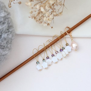 Puede incluir: Marcadores de punto hechos a mano para tejer, con aros dorados, abalorios blancos con forma de flor, pequeñas cuentas y una perla grande. Los marcadores se muestran en una aguja de tejer de madera sobre un fondo suave y neutro.