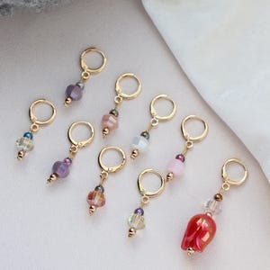May include: A selection of gold-coloured stitch markers, each with a different arrangement of colourful beads and charms. The markers feature a variety of colours, including purple, pink, and red, and are designed for use in knitting or crochet projects.