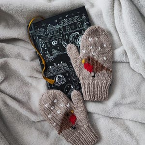 May include: A pair of knitted mittens in a light brown color, featuring a red-breasted bird design. The mittens have white heart-shaped accents and are placed on a soft, gray blanket next to a black notebook with a gold ribbon.