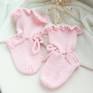 May include: A pair of light pink knitted baby mittens featuring a ruffled cuff design and a matching knitted drawstring tie at the wrist. The mittens are constructed with a simple stockinette stitch pattern and have a rounded shape at the fingertips.