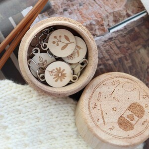 May include: A wooden box with a lid, containing several small, round wooden stitch markers with floral designs. The box is open, revealing the markers, and sits on a white knitted surface. Knitting needles are visible.