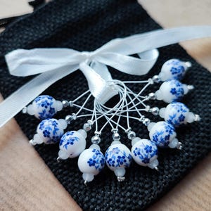 Op de afbeelding: Een set van tien witte en blauwe bloemenkralen steekmarkeerders met zilveren accenten, vastgebonden met een wit lint op een zwarte stoffen zakje. De kralen hebben een delicaat blauw bloemmotief.