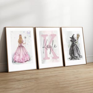 May include: Three framed art prints leaning against a wall. The prints feature a princess in a pink dress, the name "Kayla" with a large pink "K", and a witch in a black dress holding a broom. The frames are gold.