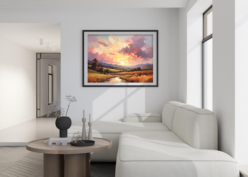 May include: A framed landscape painting depicts a sunset over a valley with a river. The artwork features vibrant colors, including orange, yellow, pink, and purple, creating a warm and inviting scene. The painting is displayed above a white sofa.