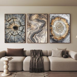Op de afbeelding: Drie ingelijste kunstdrukken met houtnerf. De prints tonen close-up weergaven van boomringen en houtnerfpatronen in tinten bruin, grijs en beige. Het kunstwerk hangt boven een beige hoekbank met decoratieve kussens en een plaid.