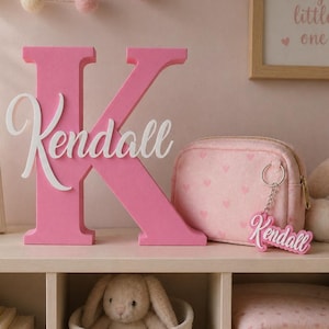 Pode incluir: Uma letra "K" rosa de madeira com o nome "Kendall" em escrita branca. Uma bolsa de cosméticos rosa com um padrão de coração e um chaveiro com o nome "Kendall" também são visíveis.